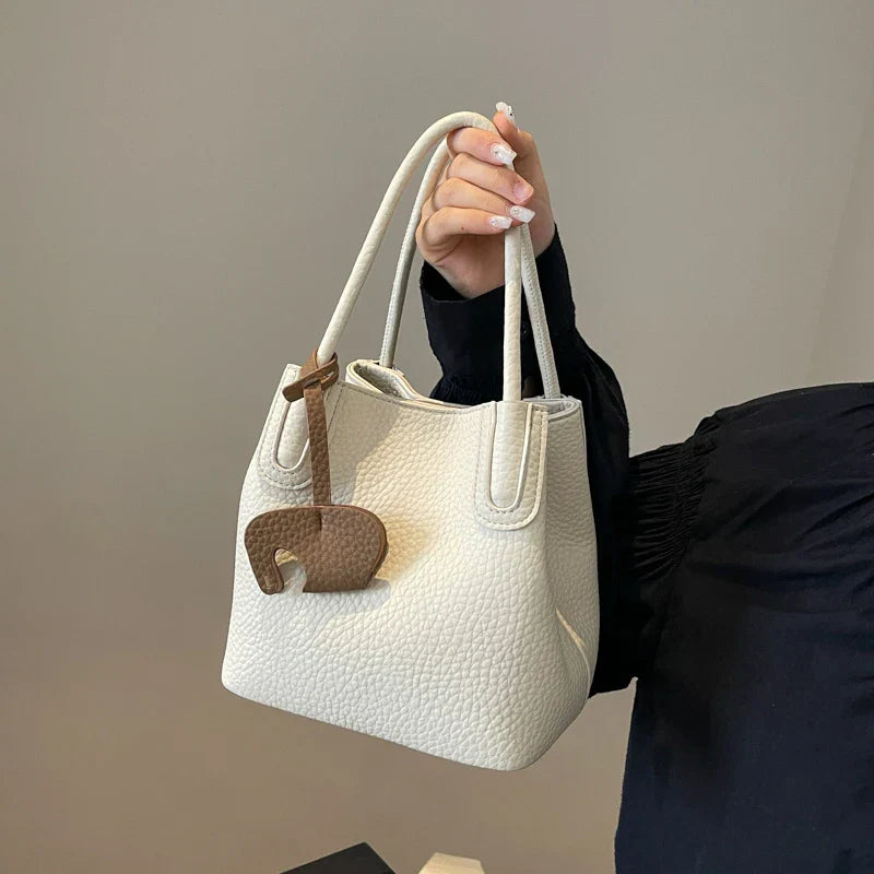 Borsa a mano in pelle bianca con texture, design elegante, manici doppi e ciondolo decorativo marrone. Accessorio moda donna di tendenza.