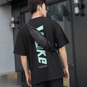 Giovane uomo con marsupio nero indossa t-shirt nera con scritta verde, all'aperto. Moda casual uomo, accessori streetwear, stile urbano.