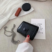 Portafoglio nero in pelle con texture coccodrillo su tavolo, accanto a catena argentata, rossetto e specchio. Accessorio moda elegante e versatile.