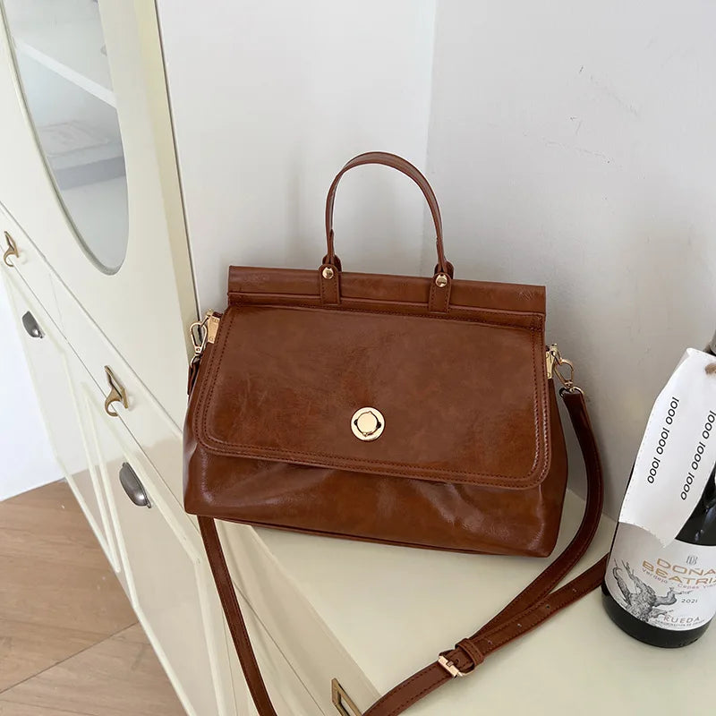 Borsa a tracolla in pelle marrone su mobile bianco, design elegante con chiusura dorata. Accessorio moda donna, stile classico e versatile.