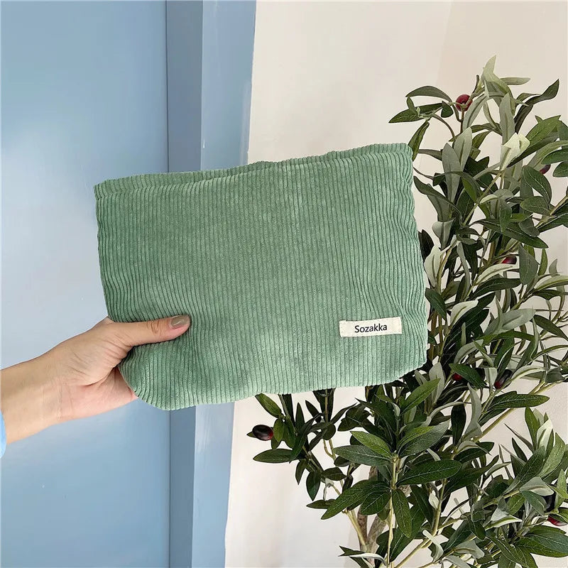 Mano che tiene una pochette verde in velluto a coste, accanto a una pianta decorativa. Accessorio moda elegante, ideale per ogni occasione.