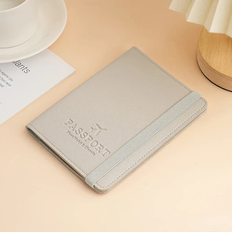 Custodia porta passaporto in pelle grigia su tavolo beige, con elastico e scritta "Passport". Accessorio da viaggio elegante e funzionale.