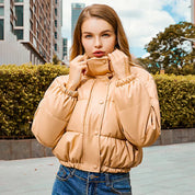Giovane donna con giacca imbottita beige, all'aperto in città. Moda autunnale, giacca calda, stile urbano. Sfondo con edifici e vegetazione.