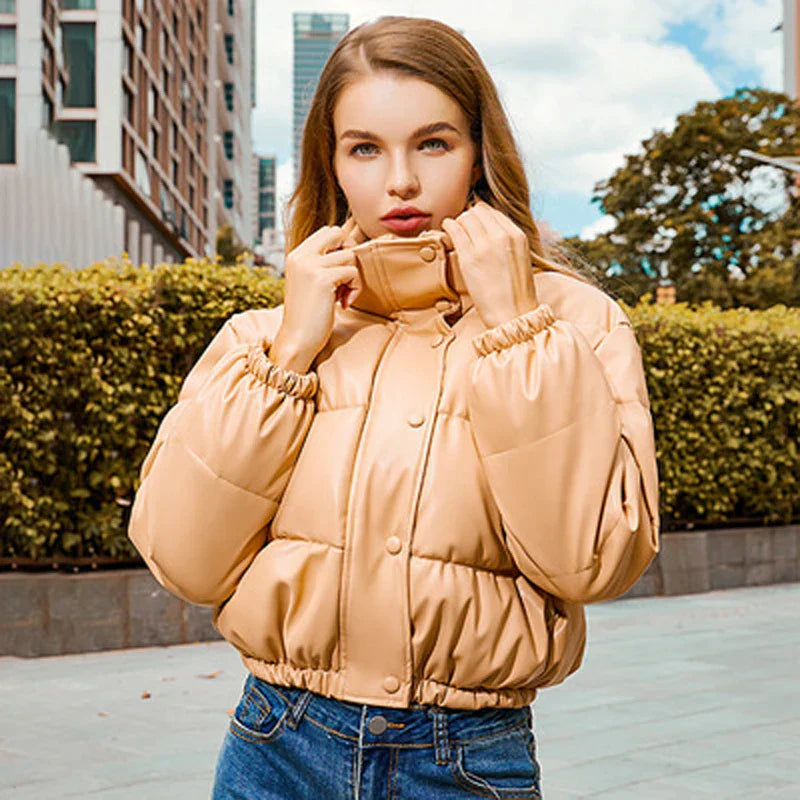Giovane donna con giacca imbottita beige, all'aperto in città. Moda autunnale, giacca calda, stile urbano. Sfondo con edifici e vegetazione.