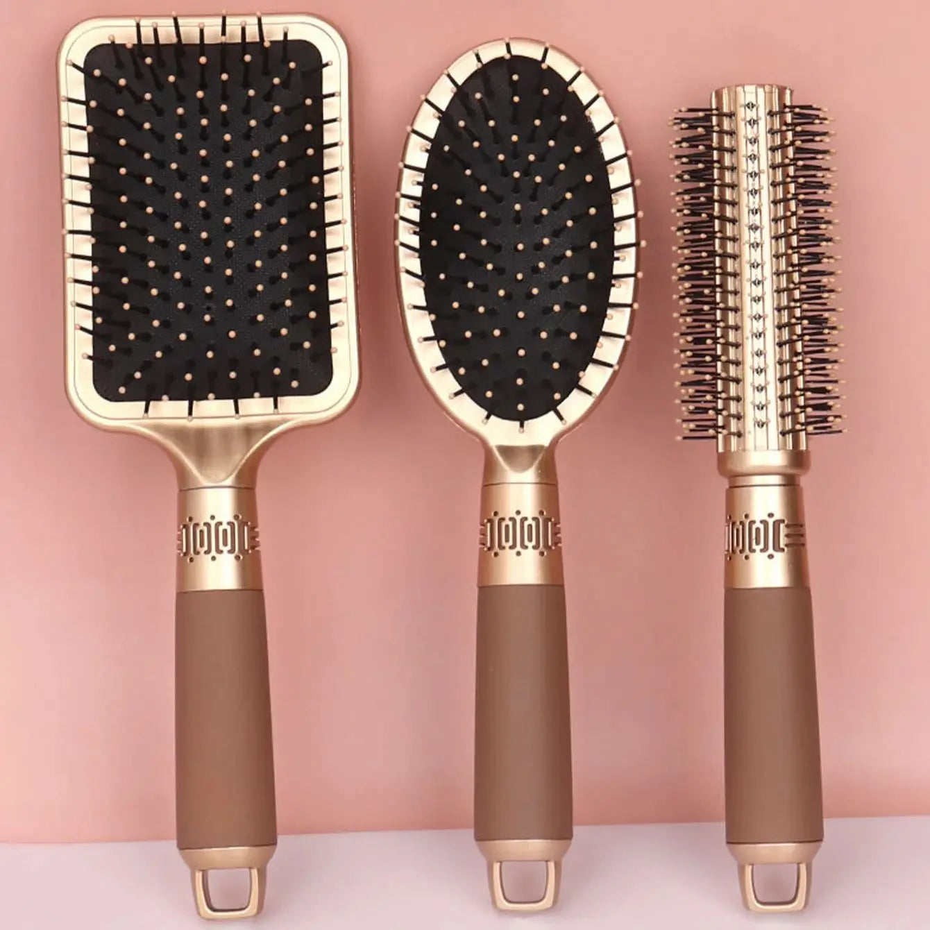 Set di tre spazzole per capelli dorate su sfondo rosa: spazzola piatta, ovale e rotonda. Accessori styling eleganti e funzionali per capelli.