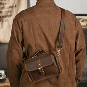 Uomo con giacca marrone indossa borsa a tracolla in pelle marrone, stile vintage, chiusura a fibbia, moda maschile, accessori eleganti.