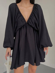 Abito nero corto con scollo a V profondo e maniche lunghe a sbuffo, tenuto da una donna. Elegante e moderno, perfetto per serate e occasioni speciali.