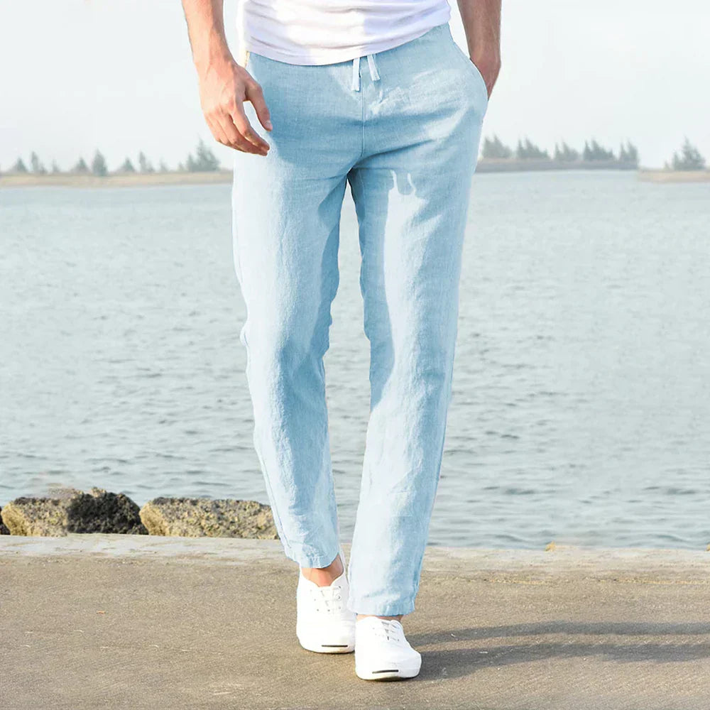 Pantaloni di lino azzurri da uomo, stile casual, con coulisse in vita, indossati con scarpe bianche, sfondo marino.