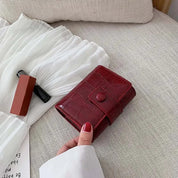 Portafoglio rosso in pelle con motivo coccodrillo su letto beige, accanto a rossetto e abito bianco. Accessorio moda elegante e funzionale.