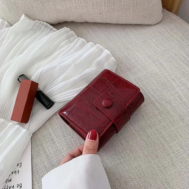Portafoglio rosso in pelle con motivo coccodrillo su letto beige, accanto a rossetto e abito bianco. Accessorio moda elegante e funzionale.