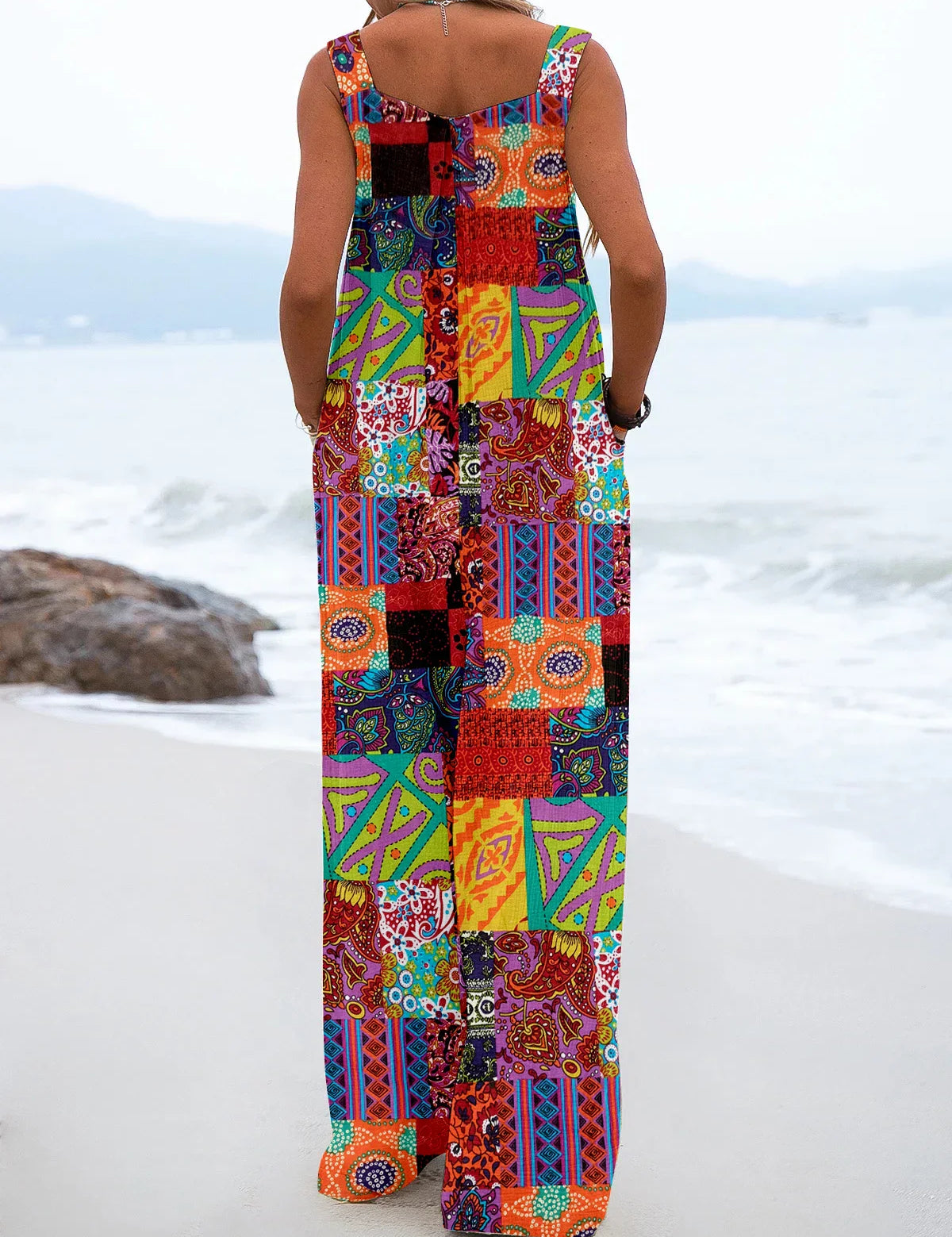 Donna in spiaggia con tuta lunga patchwork colorata, design geometrico e floreale, moda estiva, abbigliamento casual, mare sullo sfondo.