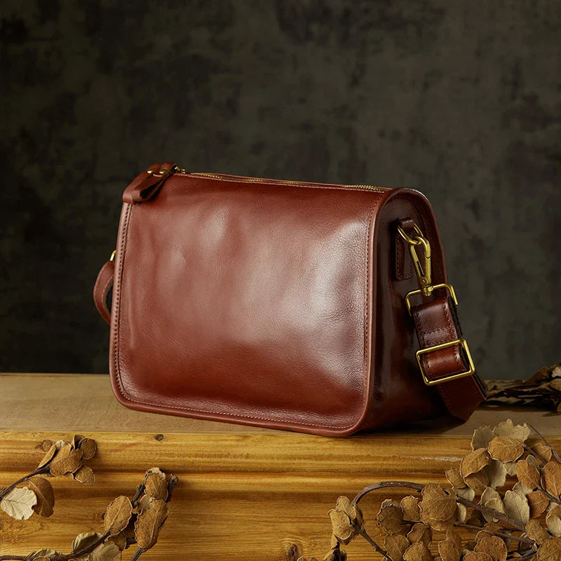 Borsa a tracolla in pelle marrone su tavolo di legno, design elegante e classico, accessorio moda uomo e donna, chiusura con fibbia dorata.