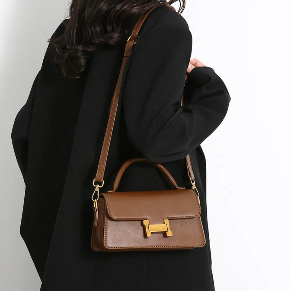 Borsa a tracolla in pelle marrone con chiusura dorata, indossata su cappotto nero. Accessorio moda elegante e versatile per donna.