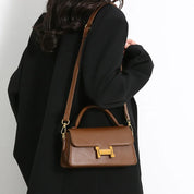 Borsa a tracolla in pelle marrone con chiusura dorata, indossata su cappotto nero. Accessorio moda elegante e versatile per donna.