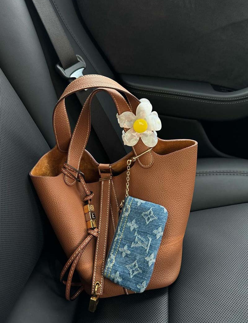Borsa in pelle marrone su sedile auto, decorata con portachiavi fiore e portafoglio in denim con motivo monogramma. Accessori moda eleganti.