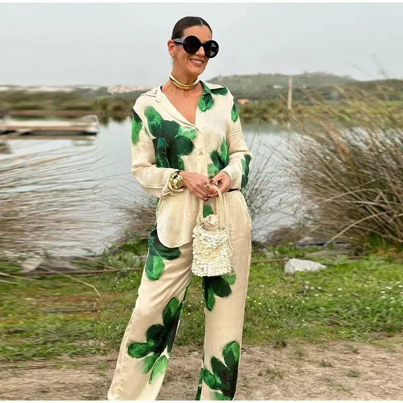 Donna con completo elegante a fantasia verde e bianco, occhiali da sole grandi, borsa a mano intrecciata, collana dorata, in ambiente naturale.