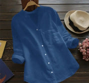 Camicia blu da donna in lino, maniche lunghe, colletto alla coreana, bottoni bianchi, su sfondo di legno con cappello di paglia e fiori rosa.