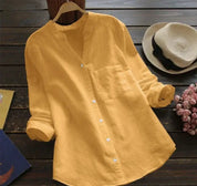 Camicia gialla in lino con colletto alla coreana, maniche lunghe arrotolate, appesa su gruccia. Sfondo con cappello di paglia e fiori rosa.