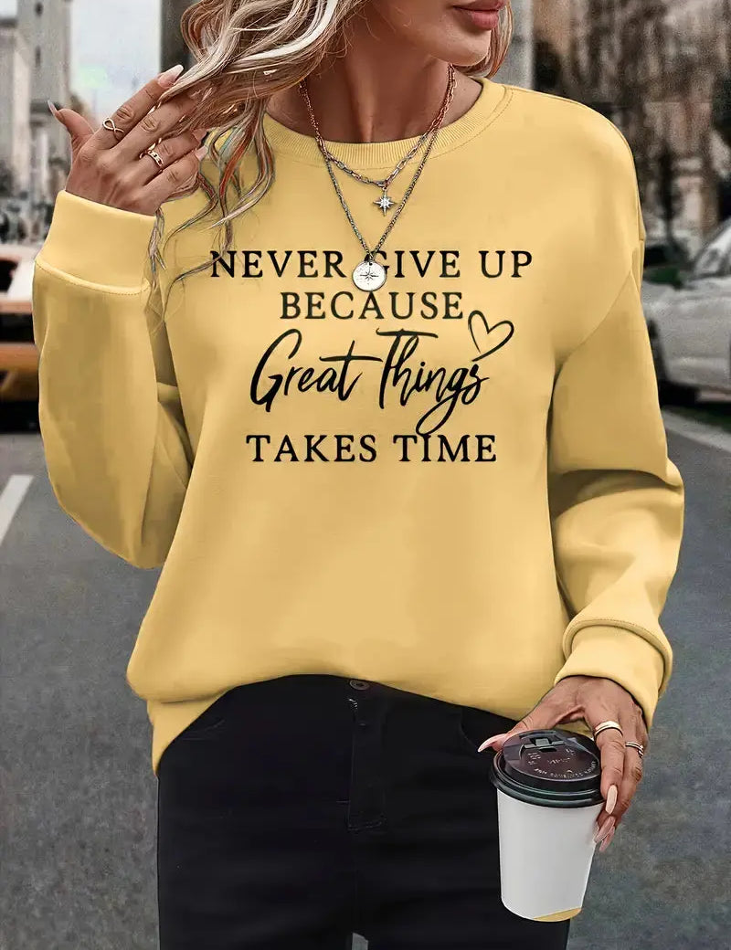 Donna con felpa gialla con scritta motivazionale, collane a strati, pantaloni neri, tiene bicchiere di caffè. Moda casual autunnale urbana.