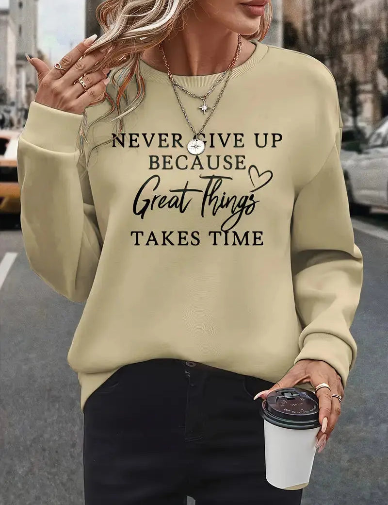 Donna con maglione beige con scritta motivazionale, collane a strati, pantaloni neri, tiene una tazza da caffè. Moda casual autunnale urbana.