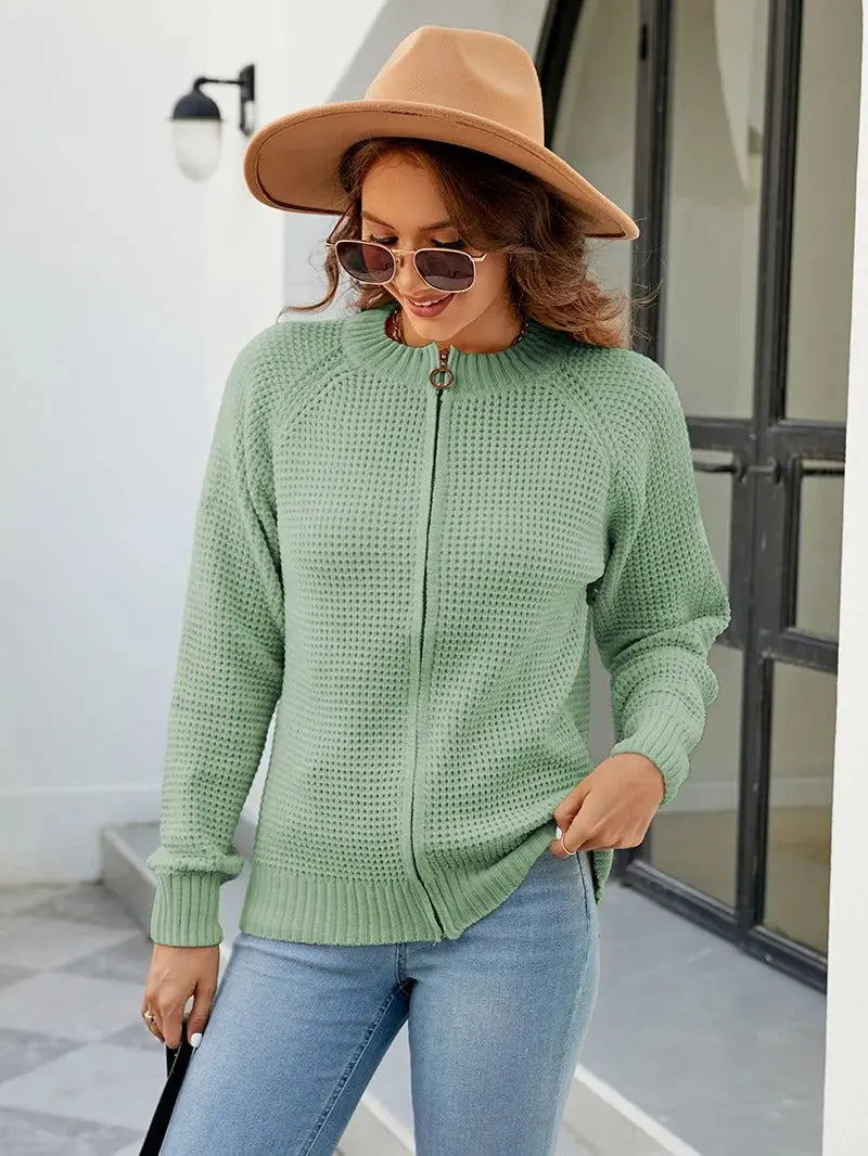 Donna con maglione verde a maglia, cappello beige e occhiali da sole, in piedi all'aperto. Moda autunnale, abbigliamento casual elegante.