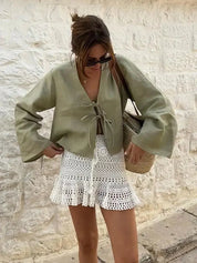 Donna con top verde oliva e gonna bianca all'uncinetto, borsa di paglia, occhiali da sole, sfondo di muro in pietra. Moda estiva casual chic.