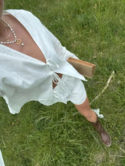 Donna in abito bianco estivo su prato verde, indossa stivali marroni e collana di perle, tiene un libro. Moda estiva, abbigliamento casual chic.