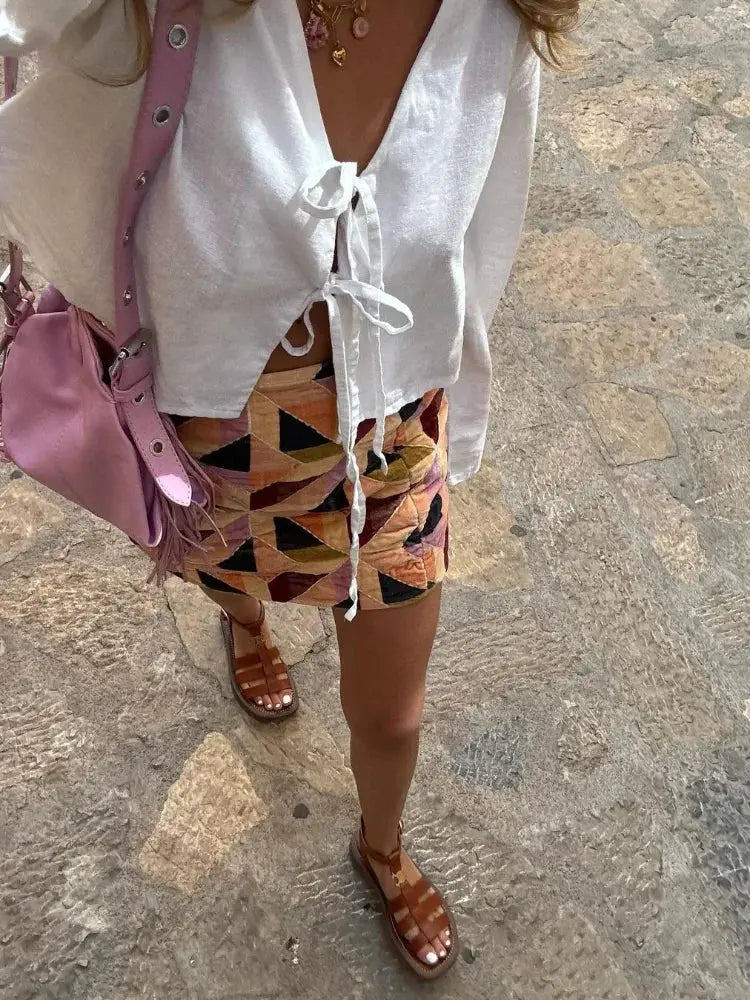 Donna con camicetta bianca annodata, gonna geometrica colorata, sandali marroni e borsa rosa su strada in pietra. Moda estiva casual.