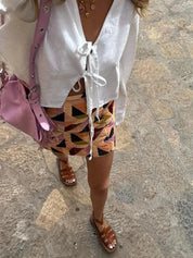 Donna con camicetta bianca annodata, gonna geometrica colorata, sandali marroni e borsa rosa su strada in pietra. Moda estiva casual.