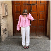 Donna con camicia rosa e pantaloni bianchi davanti a porta in legno. Indossa occhiali da sole e sandali neri. Moda estiva casual, street style.