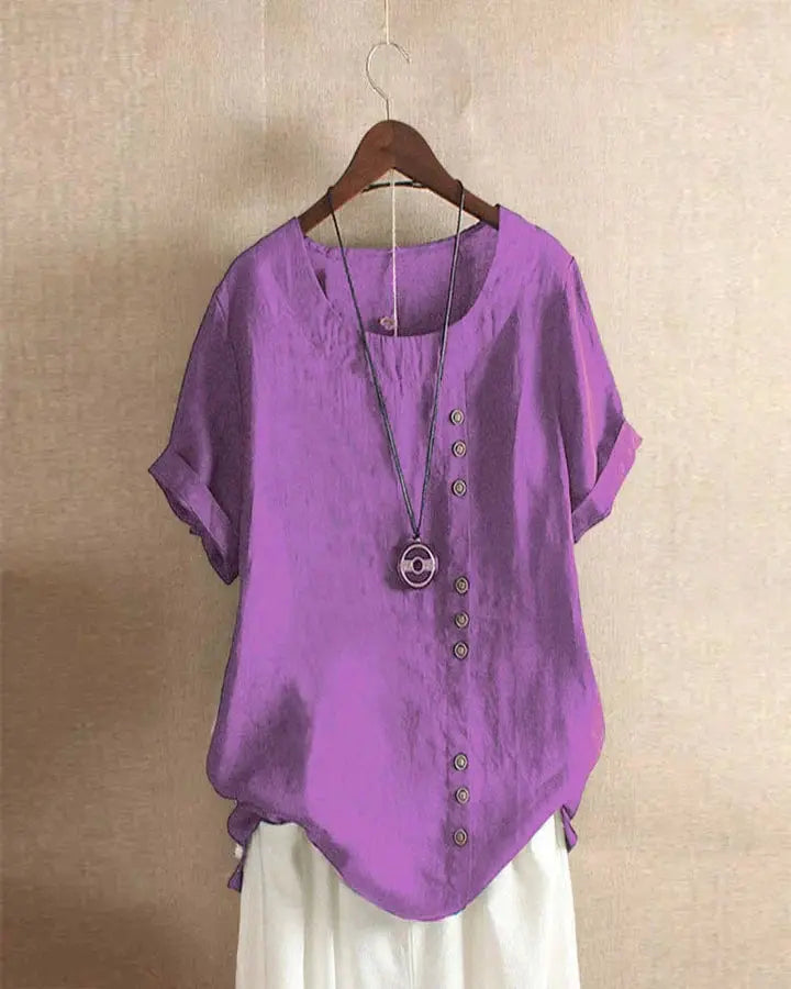 Blusa viola in lino con bottoni decorativi, maniche corte e scollo rotondo, appesa su gruccia. Abbigliamento estivo casual donna.