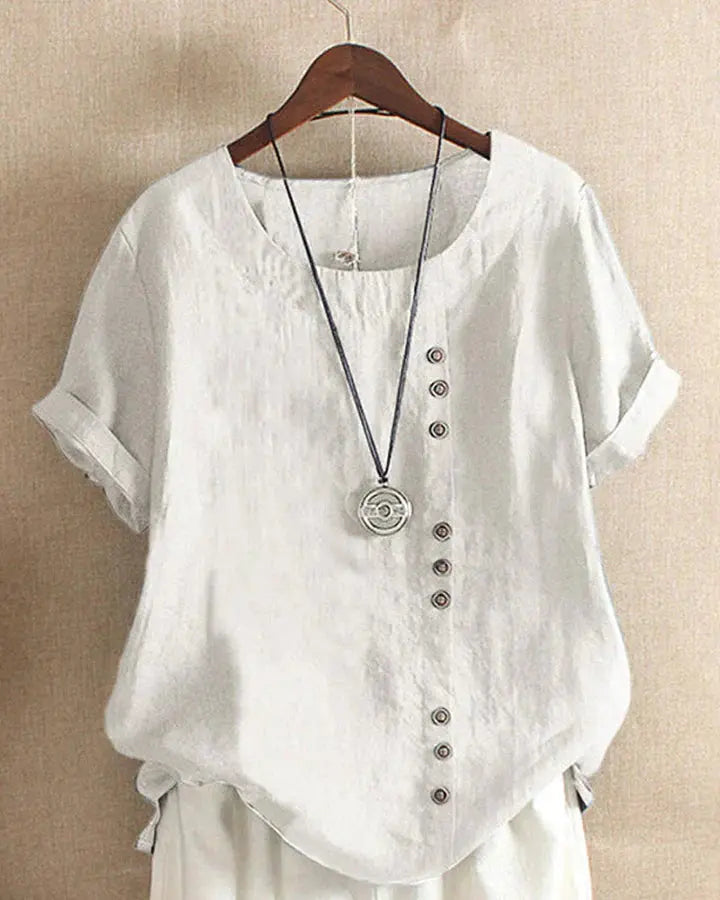 Maglietta bianca in lino con bottoni decorativi, maniche corte arrotolate e collana pendente, appesa su gruccia di legno. Moda estiva casual.