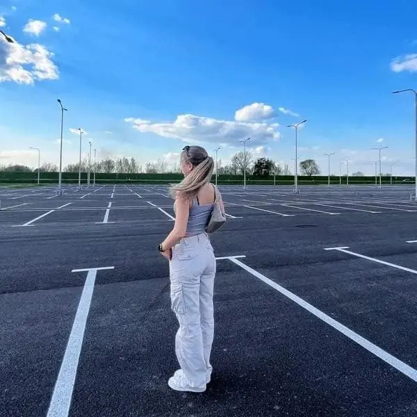 Donna in parcheggio vuoto sotto cielo blu, indossa top grigio e pantaloni bianchi cargo. Parcheggio spazioso, giornata soleggiata.