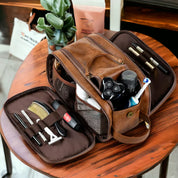 Borsa da toilette in pelle marrone aperta su tavolo, contenente articoli da toeletta come rasoio, spazzolino, pettine e flaconi. Accessori da viaggio uomo.