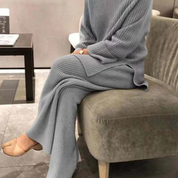 Donna seduta su divano indossa completo maglia e pantaloni grigio chiaro, tessuto a coste, moda autunnale, stile casual elegante, scarpe beige.