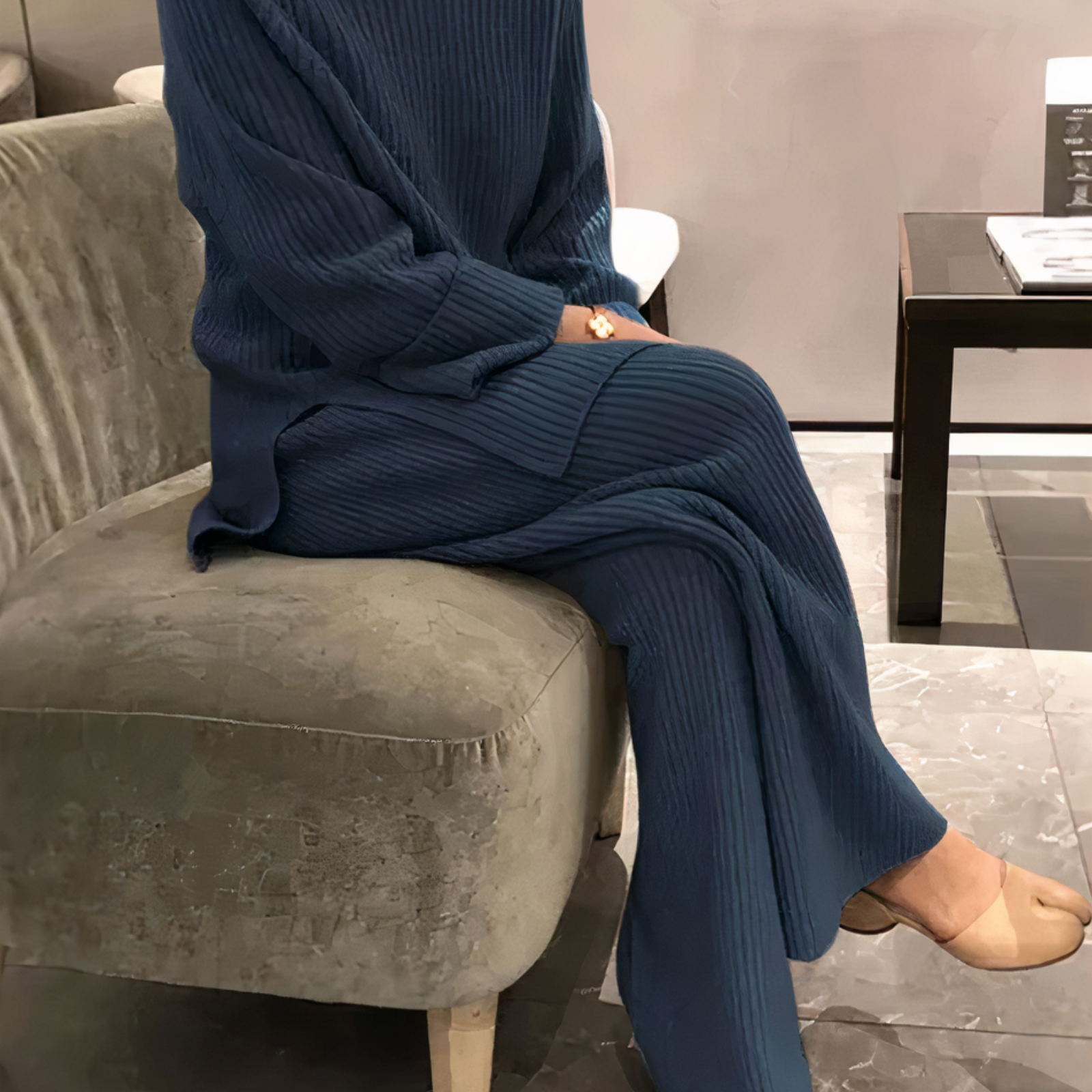Donna seduta su divano grigio indossa abito lungo blu a coste, sandali beige. Interno moderno, tavolino nero con rivista. Moda autunnale elegante.