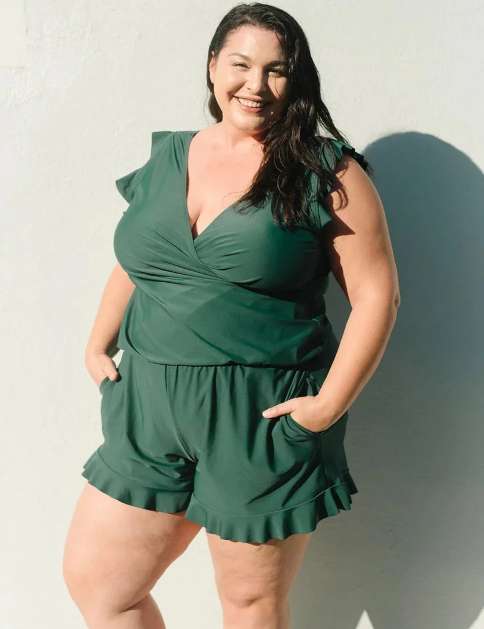 Donna sorridente in tuta verde, moda taglie forti, posa contro un muro bianco. Abbigliamento estivo elegante, stile casual chic.