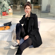 Donna seduta all'aperto con cappotto nero, jeans e sneakers Converse nere. Accanto a lei, una borsa e una scatola con baguette.