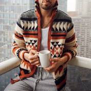 Uomo con maglione a motivi geometrici, tazza bianca in mano, su balcone cittadino. Moda autunnale, abbigliamento casual, comfort urbano.