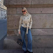 Donna con trench beige, jeans a zampa, occhiali da sole e borsa nera Chanel, appoggiata a un muro di pietra in città. Moda autunnale urbana.
