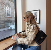 Donna bionda con occhiali da sole, giacca beige e borsa nera Chanel, seduta in un caffè vicino a una finestra, sorseggiando una bevanda calda.