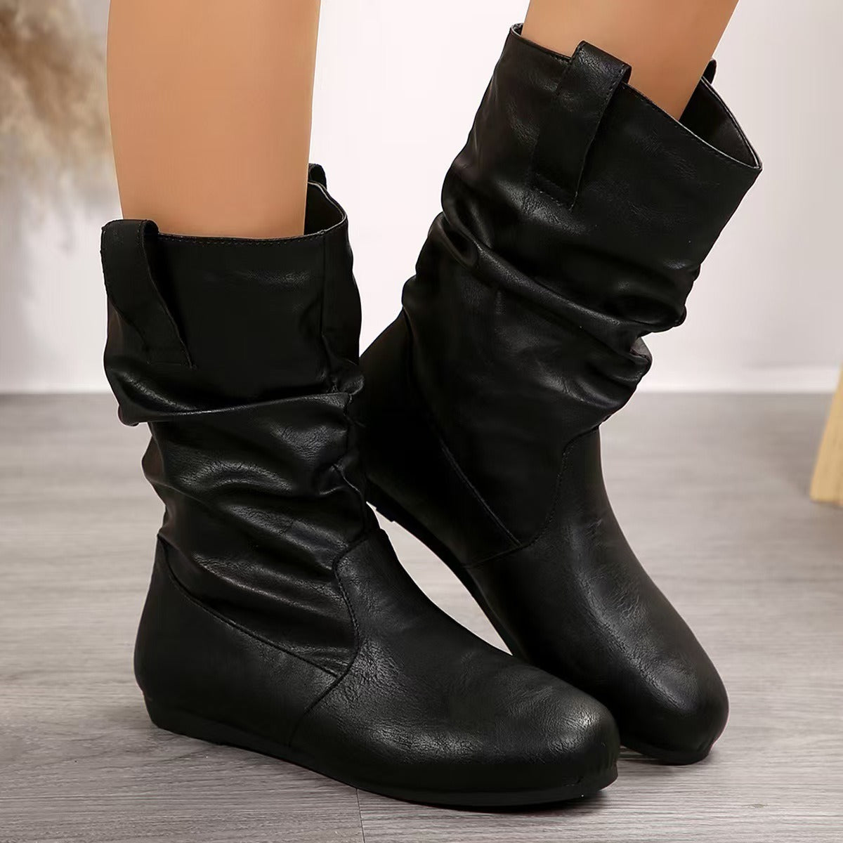 Stivali neri in pelle sintetica, design casual con pieghe, su pavimento in legno. Scarpe moda autunno inverno, comode e versatili per donna.