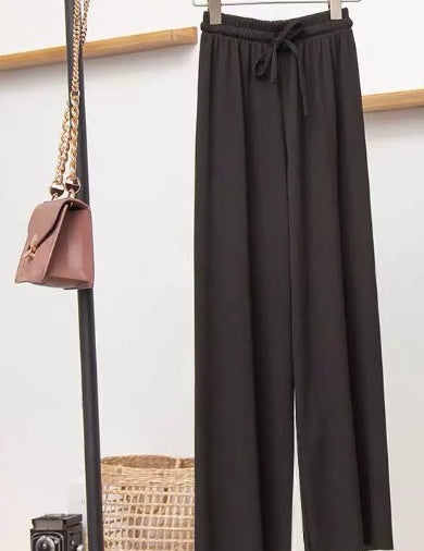 Pantaloni neri larghi appesi, borsa a tracolla marrone con catena dorata, fotocamera vintage e cesto intrecciato sullo sfondo.