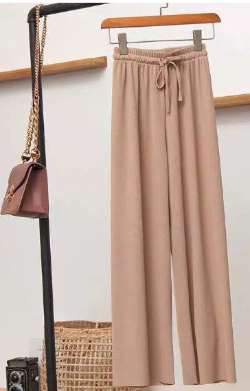 Pantaloni beige a vita alta con cintura, appesi su appendiabiti, accanto a borsa marrone con catena dorata. Moda casual elegante.