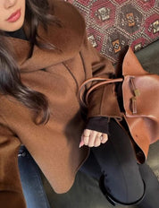 Donna con cappotto marrone elegante e borsa in pelle su sfondo di tappeto decorativo. Moda autunno inverno, accessori di lusso, stile sofisticato.