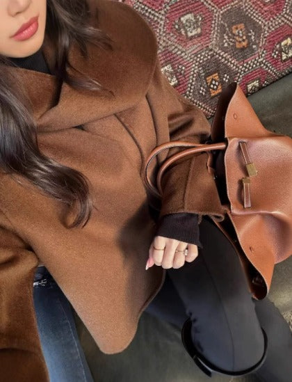 Donna con cappotto marrone elegante e borsa in pelle su sfondo di tappeto decorativo. Moda autunno inverno, accessori di lusso, stile sofisticato.