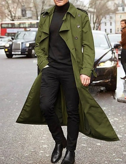 Uomo con trench verde oliva, maglione nero e pantaloni scuri, cammina in strada cittadina. Moda autunnale, stile urbano, cappotto elegante.