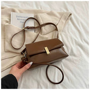 Borsa a tracolla marrone in pelle su coperta beige, con libro "Differently" accanto. Elegante accessorio moda femminile, stile casual chic.