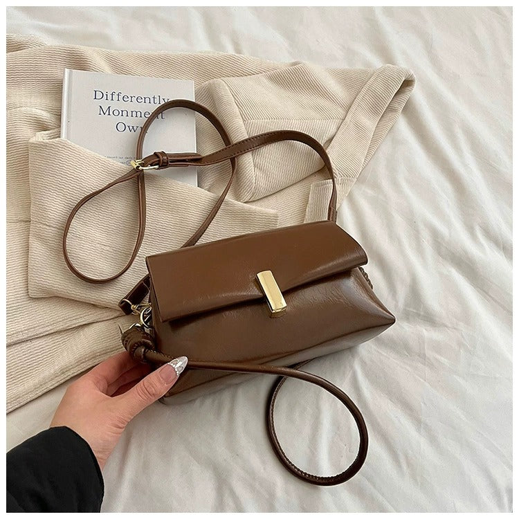 Borsa a tracolla marrone in pelle su coperta beige, con libro "Differently" accanto. Elegante accessorio moda femminile, stile casual chic.
