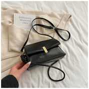 Borsa a tracolla nera in pelle su coperta beige, accanto a libro bianco. Design elegante, chiusura dorata. Accessorio moda femminile.